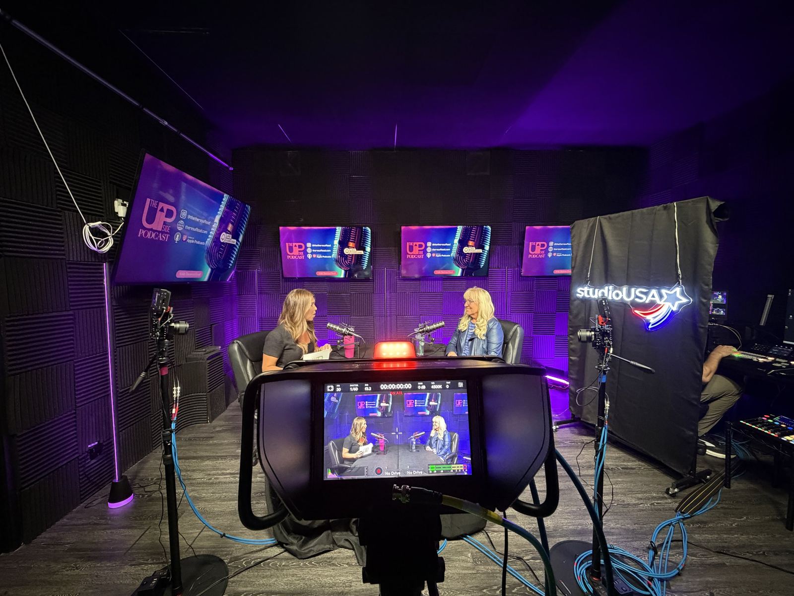 Camera viewfinder framing a two-person podcast on the purple set at studioUSA East